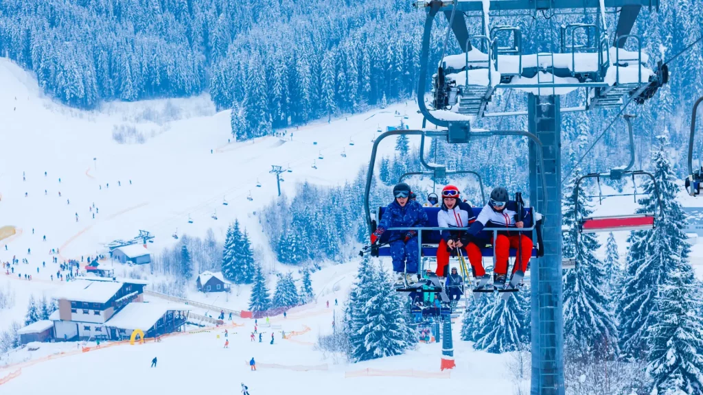 Skiers riding a chairlift above a snowy, tree-lined ski resort