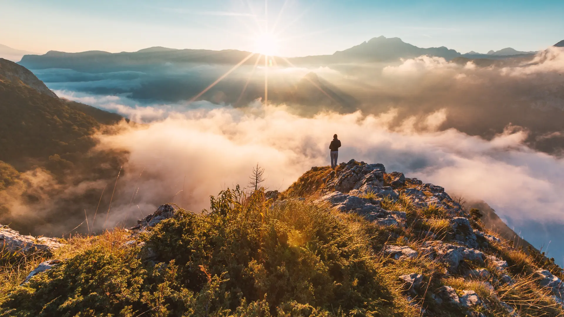 A lone figure stands on a mountain ridge above a sea of clouds at sunrise